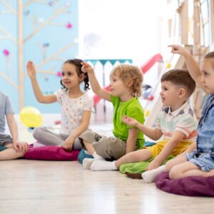 A group of children sitting on colorful cushions, raising their hands in a classroom setting.